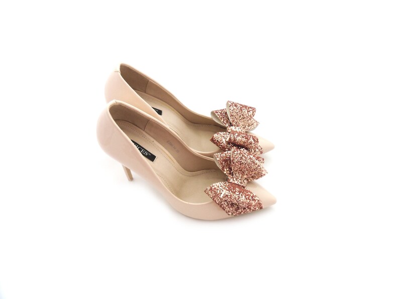 May include: A pair of nude pumps with a pointed toe and a stiletto heel. The shoes have a large, glittery bow in a rose gold color on the top of the shoe.