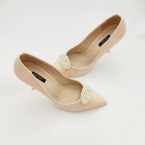 May include: A pair of nude-colored high-heeled pumps with a pearl embellishment on the toe. The shoes have a pointed toe and a stiletto heel. The brand name "VICES" is printed on the insole.