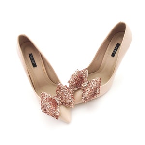 May include: A pair of nude high-heeled pumps with a rose gold glitter bow accent on each shoe. The shoes are labeled "VICES".