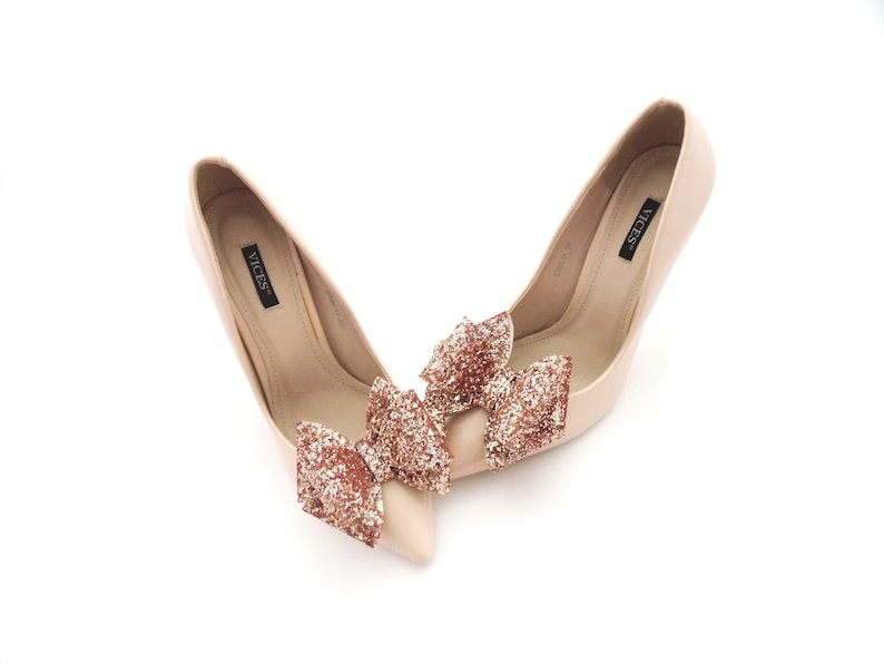 May include: A pair of nude high-heeled pumps with a rose gold glitter bow accent on each shoe. The shoes are labeled "VICES".