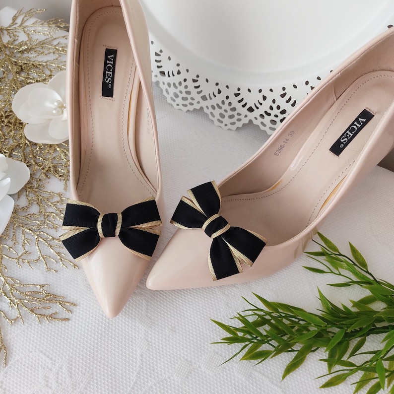 May include: A pair of nude high-heeled pumps with black and gold bows on the toes. The shoes are labeled "VICES".