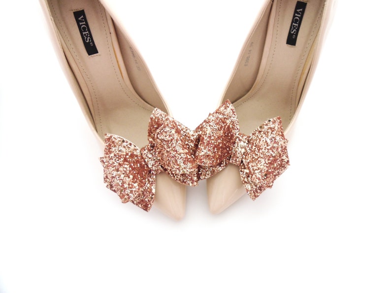 May include: A pair of nude high-heeled pumps with a rose gold glitter bow on each shoe. The shoes are labeled "VICES" on the inside.