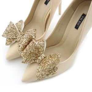 Puede incluir: Un par de zapatos de tac&oacute;n beige con punta. Cada zapato est&aacute; adornado con un gran lazo de purpurina dorada brillante. Los zapatos tienen un acabado brillante y est&aacute;n sobre un fondo blanco.