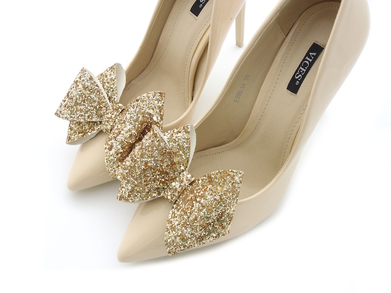 Puede incluir: Un par de zapatos de tac&oacute;n beige con punta. Cada zapato est&aacute; adornado con un gran lazo de purpurina dorada brillante. Los zapatos tienen un acabado brillante y est&aacute;n sobre un fondo blanco.