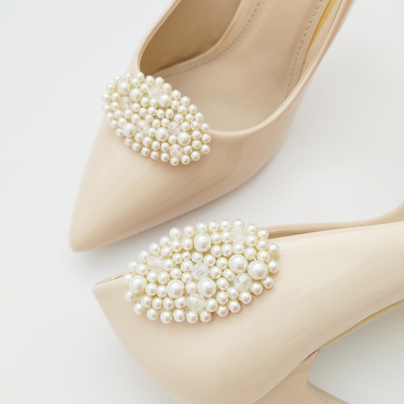 May include: A pair of beige high-heeled shoes with pearl embellishments. The shoes have a pointed toe and a delicate strap around the ankle.