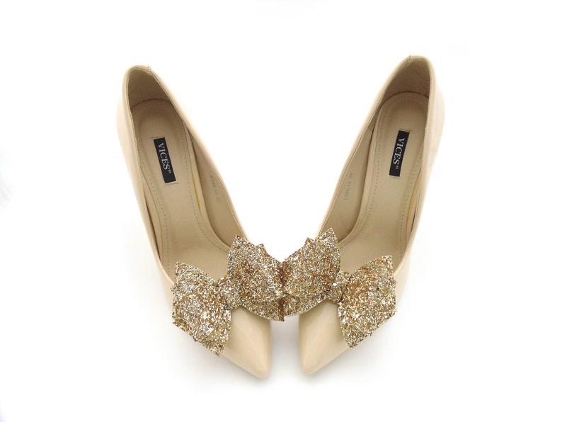 Puede incluir: Un par de tacones altos beige con punta, cada uno adornado con un gran lazo dorado brillante. Los tacones tienen la marca "VICES" impresa en la plantilla. Los zapatos est&aacute;n sobre un fondo blanco.