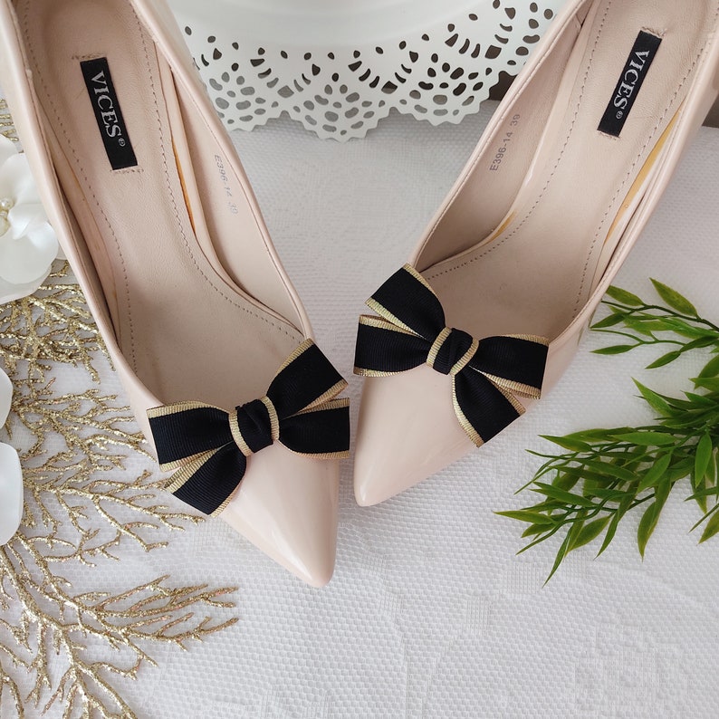 May include: A pair of nude-colored high heels with black and gold bows on the toes. The shoes are labeled "VICES" on the inside.