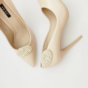 May include: A pair of nude high-heeled pumps with a pearl embellishment on the toe. The shoes are made of a smooth, shiny material and have a pointed toe.