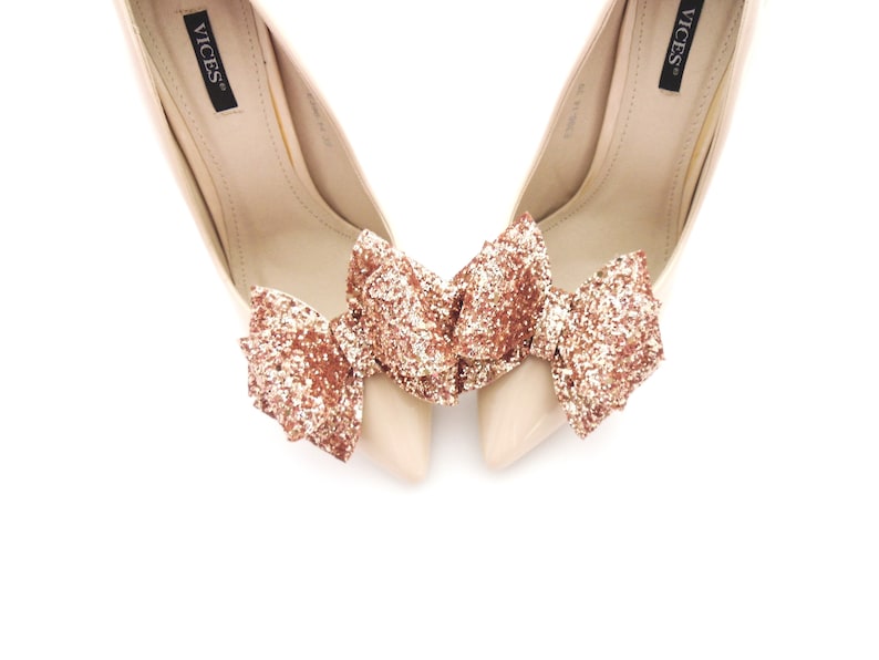 May include: A pair of nude high-heeled shoes with a rose gold glitter bow on each shoe.