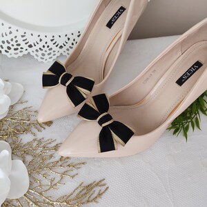May include: A pair of nude high-heeled pumps with black and gold bows on the toes. The shoes are labeled "VICES" on the side.