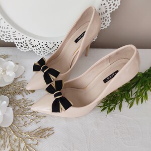 May include: A pair of nude high-heeled pumps with black and gold bows on the toes. The shoes are labeled "VICES".