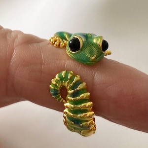 May include: A gold-toned open ring shaped like a gecko, featuring vibrant green and blue enamel detailing on the body and head. The lizard has prominent black eyes and textured gold accents along its back, tail, and feet, creating a wrap-around design.