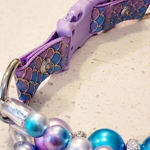 May include: A purple and blue patterned dog collar with a silver buckle and a matching beaded charm. The beads are iridescent and have a pearl-like finish.