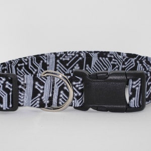 May include: A black and white dog collar with a circuit board pattern. The collar has a black plastic buckle and a silver metal D-ring for leash attachment. The collar is designed for pets.