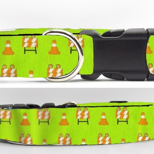 Puede incluir: Un collar de perro verde con un patrón repetido de conos de tráfico naranja y señales de tráfico blancas y naranjas. El collar tiene una hebilla de plástico negra y un anillo en D de metal plateado.