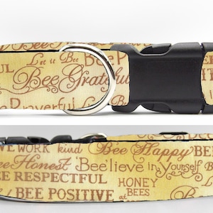 May include: A yellow dog collar with a black buckle and a silver ring. The collar is printed with the words "Bee Happy", "Bee Honest", "Bee Respectful", "Bee Positive", and other bee-themed phrases in brown lettering.