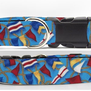 May include: A blue dog collar with a nautical pattern of red, white, and blue flags, yellow anchors, and gold rope. The collar has a black buckle and a silver D-ring.