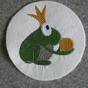 Könnte beinhalten: Gestickter Aufnäher mit einer grünen Kröte, die eine goldene Krone trägt und eine goldene Kugel hält. Die Kröte befindet sich auf einem weißen Hintergrund.