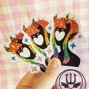 May include: Three iridescent rainbow glitter stickers featuring a black silhouette of a demon with red hair and horns. The demon has a heart-shaped face with a white outline. The stickers have a white background and are decorated with blue stars. The stickers are held in a hand and a sticker with the text "nena mágica" is visible in the background.