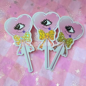May include: Three heart-shaped stickers with a cartoon eye design, a gold glitter bow, and a lollipop stick. The hearts are pink with a white outline. The background is a pink and white checkered pattern.