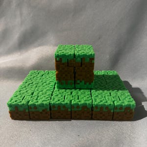 May include: A collection of green and brown pixelated blocks, resembling a grassy surface and a stacked cube. The blocks are arranged to create a landscape scene, reminiscent of a video game environment.