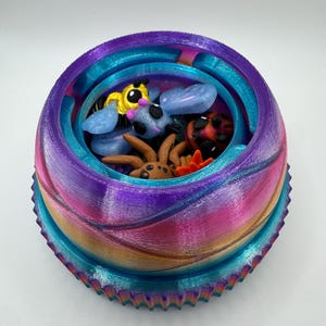 Puede incluir: Un contenedor circular, colorido y en capas con una variedad de pequeñas figuras de insectos de dibujos animados en el interior. El contenedor tiene un exterior texturizado y acanalado con un degradado de colores, incluyendo morado, rosa y turquesa. Los insectos incluyen una abeja, una mariquita y una araña.