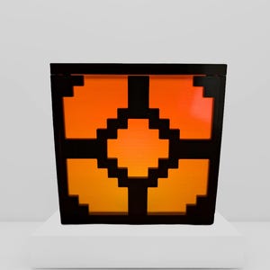 Pode incluir: Uma caixa quadrada preta com um design pixelizado laranja e amarelo. O design é um padrão geométrico de quadrados e retângulos. A caixa está sobre uma superfície branca. A caixa tem aproximadamente 20 cm de largura.