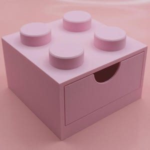 Może przedstawiać: Różowe pudełko do przechowywania zaprojektowane tak, aby przypominało klocek konstrukcyjny, z czterema podniesionymi okrągłymi kołkami na górze. Pudełko ma jedną szufladę z zakrzywionym uchwytem, ​​wszystko w pasującym różowym odcieniu.