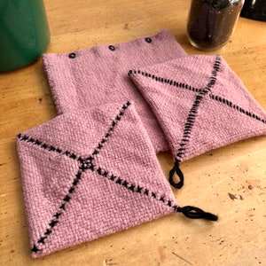 May include: Three pink knitted pot holders with black stitching. The pot holders have a diamond pattern with a black X in the centre. The pot holders have black tassels.