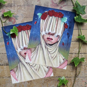 May include: Two art prints featuring a surreal portrait. The subject has a cream-colored veil with vertical folds, red roses, and a blue background with falling red petals. The prints are on a wooden surface.