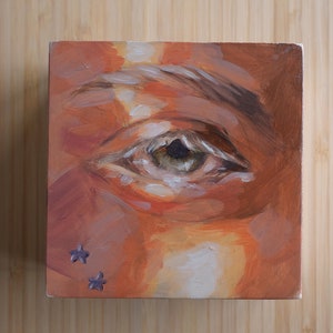 May include: Close-up painting of a human eye with a brown iris and a light brown eyebrow. Two small purple stars are painted on the lower eyelid.