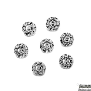 Może przedstawiać: Siedem srebrnych metalowych guzików z ozdobnym wzorem i literą w środku. Litery to A, H, K, R, S, F i X.