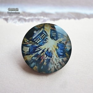 Puede incluir: Un cabujón redondo con una representación artística azul y amarilla de una cabina de policía, que recuerda a un tema de ciencia ficción. El cabujón tiene un acabado brillante y está sobre un fondo neutro. El texto "CHDM Cabochon" es visible.