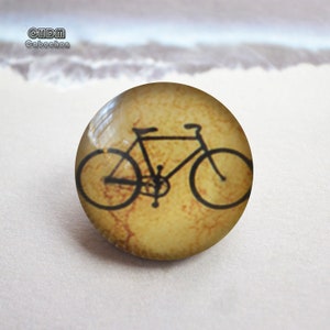 May include: A round cabochon with a bicycle illustration. The bicycle is black and the background is a mottled tan color. The cabochon has a glossy finish. The text "CHDM Cabochon" is in the upper left corner.
