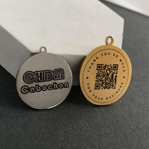 Puede incluir: Dos colgantes redondos de metal. Uno es plateado con el texto "CHDM Cabochon". El otro es dorado con un código QR y el texto "THANK YOU SO MUCH FOR YOUR BUSINESS" en el borde. Ambos tienen un pequeño lazo en la parte superior.