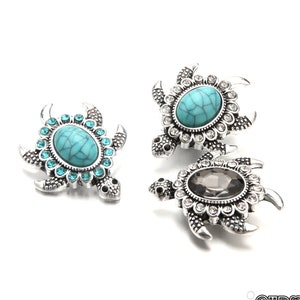 以下が含まれることがあります： 銀色のカメの形をした3つのジュエリー。2つのカメは、透明なクリスタルに囲まれたターコイズ色の楕円形の中心を持っています。3つ目は、透明なクリスタルにも囲まれた灰色の楕円形の中心を持っています。カメは、詳細なヒレと頭のデザインをしています。