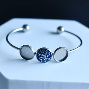 May include: A silver-toned cuff bracelet with three circular settings. The center setting features a sparkling blue and green druzy stone, while the other two settings are empty. The bracelet has a simple, elegant design.