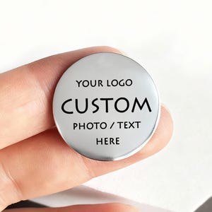 Op de afbeelding: Een ronde, zilverkleurige metalen schijf met zwarte gegraveerde tekst die luidt YOUR LOGO CUSTOM PHOTO / TEXT HERE. Het oppervlak is glad en reflecterend, wat zorgt voor een strakke, metalen afwerking voor persoonlijke aanpassingen.