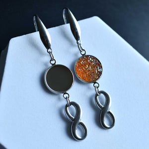 May include: Silver-toned dangle earrings. One earring features a dark gray circular pendant, while the other has a sparkling orange and gold circular pendant. Both earrings have an infinity symbol charm.