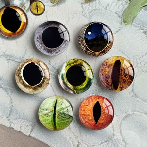 May include: A collection of glass eye cabochons in various colors and designs. The eyes feature a black pupil and iris patterns in shades of yellow, gray, brown, green, and orange. Some eyes are set in metal pendants.