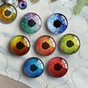 May include: A collection of glass eye cabochons in various colors, including yellow, orange, blue, green, and red. Some have glitter effects. One is set in a silver pendant. The irises have a detailed, realistic appearance.