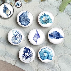 May include: A collection of round, white-backed pendants and magnets featuring watercolor illustrations of various seashells in shades of blue. The designs include spiral and conch shell shapes, perfect for coastal decor.