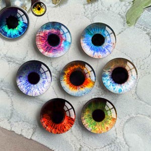 May include: A selection of glass eye cabochons in diverse colours, such as blue, green, red, and orange. Each eye features a black pupil and detailed, colourful patterns. One eye is set in a silver pendant. Ideal for jewellery making or crafting projects.