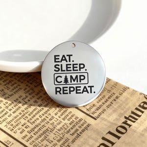 May include: A circular, silver-toned pendant featuring the text "EAT. SLEEP. CAMP. REPEAT." in black. The word "CAMP" is within a rectangle with a tree symbol. The pendant has a small hole at the top for attachment.