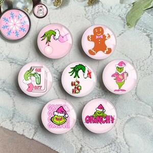 May include: A selection of pink and clear glass cabochons featuring festive Christmas designs. The designs include a gingerbread man, a snowflake, and the Grinch in various poses. The cabochons are round and have a glossy finish.