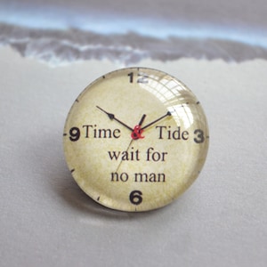 以下が含まれることがあります： 「Time & Tide wait for no man」の文字が入った、ガラスで覆われた丸い時計の文字盤。 黒い針が時間を指しています。 3、6、9、12の数字が見えます。 背景はヴィンテージ風の紙のようです。
