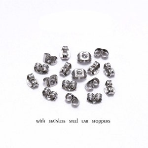May include: A close-up of a group of silver earring backs, also known as ear stoppers, scattered on a white surface.  The text "with stainless steel ear stoppers" is printed below the earring backs.