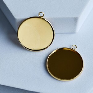 Puede incluir: Dos colgantes redondos de color dorado con un acabado pulido. Cada colgante tiene un peque&ntilde;o lazo en la parte superior para sujetarlo a una cadena u otro componente de joyer&iacute;a. Los colgantes se muestran sobre una superficie blanca.