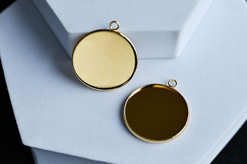 Puede incluir: Dos colgantes redondos de color dorado con un acabado pulido. Cada colgante tiene un peque&ntilde;o lazo en la parte superior para sujetarlo a una cadena u otro componente de joyer&iacute;a. Los colgantes se muestran sobre una superficie blanca.
