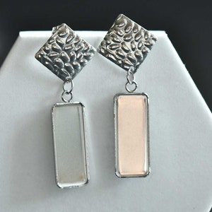May include: A pair of silver dangle earrings with a textured diamond-shaped top and a rectangular glass bottom. One earring has a clear glass bottom, and the other has a light pink glass bottom.